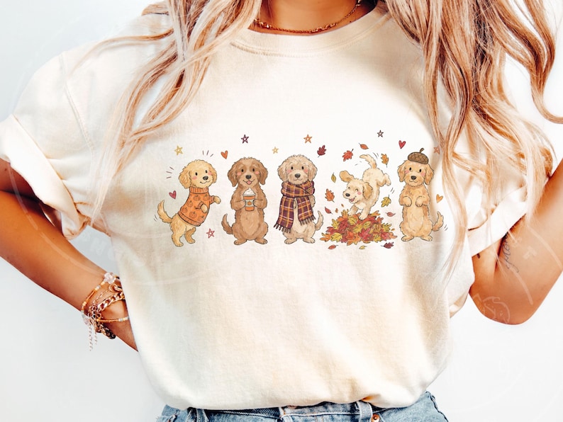 May include: A cream-colored t-shirt featuring a graphic print of five golden-colored dogs in various autumn-themed poses. The dogs are depicted wearing sweaters, scarves, and an acorn hat, with some playing in a pile of colorful fall leaves.