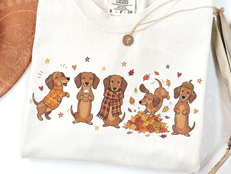 May include: A cream-colored t-shirt featuring a graphic print of five brown dachshunds in autumn-themed attire. The dogs are depicted wearing a pumpkin-patterned sweater, holding a coffee cup, wearing a plaid scarf, playing in leaves, and wearing an acorn hat.