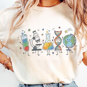 Puede incluir: Una camiseta de color crema con un estampado gr&aacute;fico colorido de equipos cient&iacute;ficos animados que incluyen un tubo de ensayo, un microscopio, un matraz, una cadena de ADN y un globo terr&aacute;queo, todos con caras sonrientes y peque&ntilde;as estrellas.