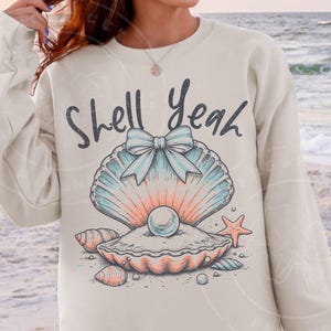 May include: Cream-colored sweatshirt featuring a seashell graphic with the text "Shell Yeah." The graphic includes a large seashell with a pearl, a bow, and other seashells and a starfish. The design is in shades of blue, peach, and gray.
