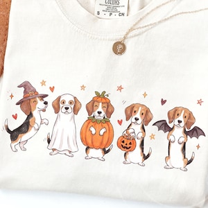 Peut inclure: Un t-shirt couleur crème arborant un motif de cinq chiens beagle déguisés pour Halloween, incluant un chapeau de sorcière, un drap de fantôme, un costume de citrouille, un seau citrouille et des ailes de chauve-souris, avec des étoiles et cœurs.