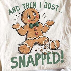 Peut inclure: Un t-shirt couleur crème avec une illustration d'un bonhomme en pain d'épices marron portant un nœud papillon vert et des boutons rouges. Le bonhomme a une jambe cassée avec des miettes. Le texte indique : AND THEN I JUST... SNAPPED!