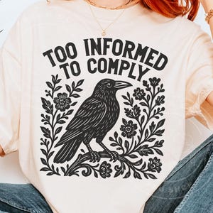 Pode incluir: Camiseta creme com um desenho gr&aacute;fico preto de um corvo empoleirado em um galho, rodeado por elementos florais. O texto "TOO INFORMED TO COMPLY" est&aacute; arqueado acima do corvo. O design &eacute; impresso em uma camiseta macia e casual.