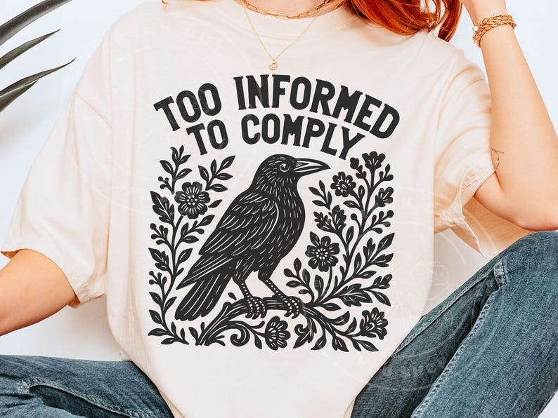Pode incluir: Camiseta creme com um desenho gr&aacute;fico preto de um corvo empoleirado em um galho, rodeado por elementos florais. O texto "TOO INFORMED TO COMPLY" est&aacute; arqueado acima do corvo. O design &eacute; impresso em uma camiseta macia e casual.