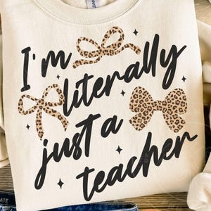 May include: Cream-colored sweatshirt with the text "I'm literally just a teacher" in black script. The design includes three leopard print bows and small black star accents, creating a playful and stylish look.