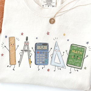 Pode incluir: Uma t-shirt de cor creme com uma impressão gráfica de cinco materiais escolares antropomórficos: uma régua de madeira, um compasso de metal, uma calculadora azul, um esquadro transparente e um livro verde com a palavra MATH na capa.