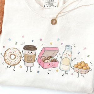 以下が含まれることがあります： クリーム色のTシャツで、ドーナツ、コーヒーカップ、ピンクのドーナツ箱、牛乳のガラス瓶、ドーナツホールが入った小さなトレイという、擬人化された5つの食べ物のグラフィックプリントが施されており、周囲には小さなカラフルな星が散りばめられています。