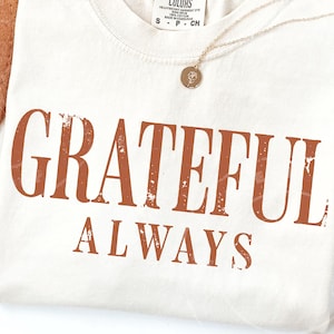 May include: A cream-colored t-shirt featuring the text GRATEFUL ALWAYS printed in a distressed, burnt orange font. The shirt has a crew neckline and a small tag at the collar. A thin gold-toned chain necklace with a small circular pendant rests on top.