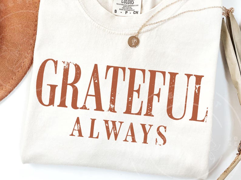 May include: A cream-coloured t-shirt featuring the text GRATEFUL ALWAYS printed in a distressed, burnt orange font. The shirt has a crew neckline and a small tag at the collar. A thin gold-toned chain necklace with a small circular pendant rests on top.
