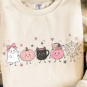 May include: A cream-colored crewneck sweatshirt featuring a graphic print of a white ghost with a pink bow, a pink pumpkin, a black cat mug, a pink candy bucket, and a spiderweb, all accented with small pink hearts and stars.