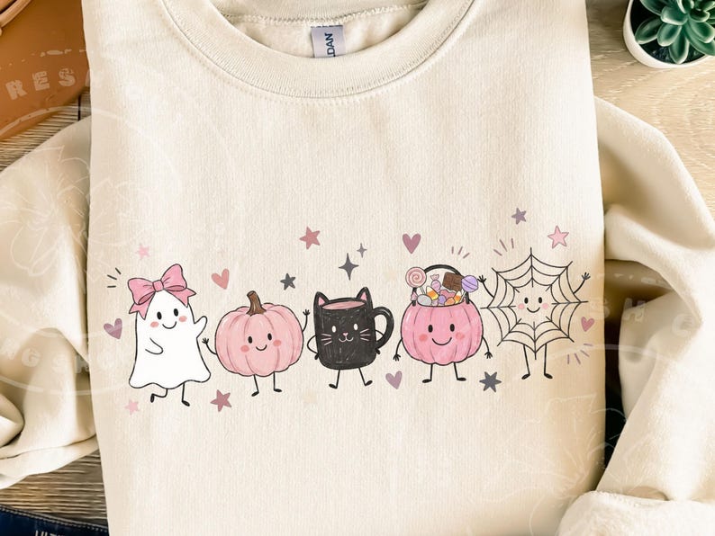 May include: A cream-colored crewneck sweatshirt featuring a graphic print of a white ghost with a pink bow, a pink pumpkin, a black cat mug, a pink candy bucket, and a spiderweb, all accented with small pink hearts and stars.
