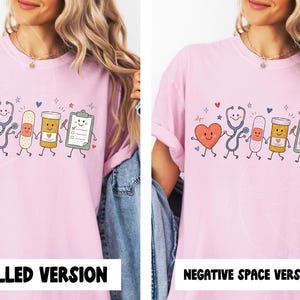May include: A light pink t-shirt featuring a graphic print of five anthropomorphic medical items holding hands: a heart, a stethoscope, a bandage, a pill bottle, and a clipboard. The design includes small colorful hearts and star accents.