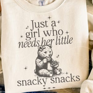 May include: Cream-colored sweatshirt with the text "Just a girl who needs her little snacky snacks." The graphic features a bear and a smaller animal. The sweatshirt is a crew neck style.