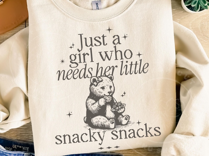 May include: Cream-colored sweatshirt with the text "Just a girl who needs her little snacky snacks." The graphic features a bear and a smaller animal. The sweatshirt is a crew neck style.