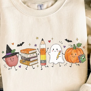 May include: A cream-colored crewneck sweatshirt featuring a colorful graphic print of a smiling apple wearing a witch hat, a stack of books with a spider web, a pencil, a ghost holding a pencil, and a pumpkin holding a small book.
