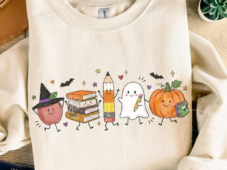 May include: A cream-colored crewneck sweatshirt featuring a colorful graphic print of a smiling apple wearing a witch hat, a stack of books with a spider web, a pencil, a ghost holding a pencil, and a pumpkin holding a small book.