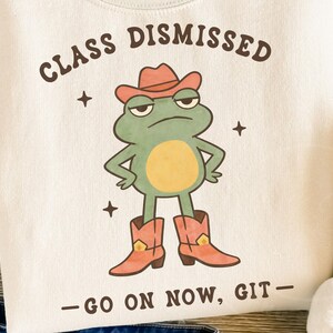 May include: Cream-colored sweatshirt featuring a cartoon frog wearing a cowboy hat and boots. The frog has a grumpy expression and is standing with its arms crossed. The text "CLASS DISMISSED - GO ON NOW, GIT-" is printed above and below the frog.
