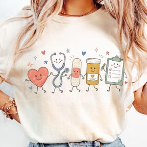 May include: A cream-colored t-shirt featuring a graphic print of five anthropomorphic medical items holding hands: a red heart, a blue stethoscope, a tan adhesive bandage, a yellow pill bottle, and a green clipboard with a checklist.