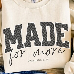 Puede incluir: Sudadera color crema con las palabras "MADE for more" en estampado de leopardo y cuadros negros y grises. El texto "Efesios 2:10" est&aacute; impreso debajo. La sudadera est&aacute; doblada.