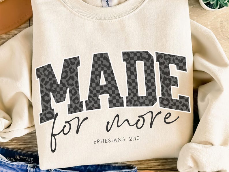 Puede incluir: Sudadera color crema con las palabras "MADE for more" en estampado de leopardo y cuadros negros y grises. El texto "Efesios 2:10" est&aacute; impreso debajo. La sudadera est&aacute; doblada.