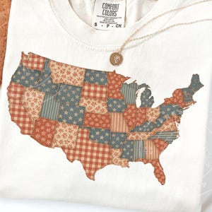 May include: Cream-colored t-shirt featuring a patchwork design of the United States map. The map is composed of various patterns in red, blue, and beige, including stars, stripes, and floral prints. A gold necklace with a pendant rests on the shirt.