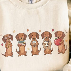 May include: A cream-colored crewneck sweatshirt featuring a graphic of five brown dachshunds. The dogs are depicted with various items including glasses and a pencil, a stack of books, an iced coffee, a clipboard, and a red apple, accented by small hearts and stars.