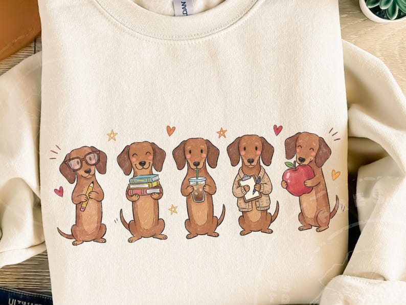 May include: A cream-colored crewneck sweatshirt featuring a graphic of five brown dachshunds. The dogs are depicted with various items including glasses and a pencil, a stack of books, an iced coffee, a clipboard, and a red apple, accented by small hearts and stars.