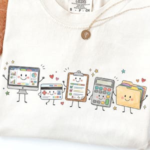 Peut inclure: Un t-shirt couleur crème avec une impression graphique représentant cinq objets de bureau anthropomorphes : un écran d'ordinateur, une carte d'identité, un presse-papiers avec une liste, une calculatrice et un dossier jaune, tous souriants.