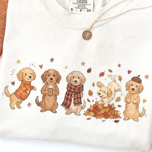 May include: A cream-colored t-shirt featuring a graphic illustration of five golden-colored dogs in autumn-themed attire and poses, including one wearing a pumpkin sweater, one holding a coffee cup, one with a plaid scarf, one playing in leaves, and one wearing an acorn hat.
