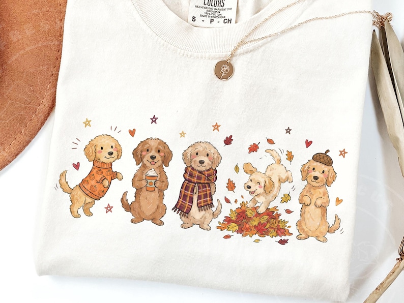 May include: A cream-colored t-shirt featuring a graphic illustration of five golden-colored dogs in autumn-themed attire and poses, including one wearing a pumpkin sweater, one holding a coffee cup, one with a plaid scarf, one playing in leaves, and one wearing an acorn hat.