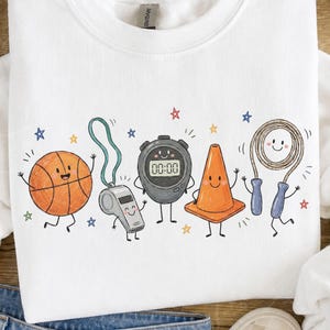 May include: A white crewneck sweatshirt featuring a graphic print of anthropomorphic sports equipment including an orange basketball, a silver whistle, a black stopwatch displaying 00:00, an orange traffic cone, and a jump rope with blue handles.
