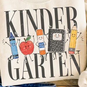 May include: A cream-colored sweatshirt featuring the text KINDER GARTEN in large dark gray letters. Between the words are cartoon illustrations of a blue crayon, a red apple, an orange glue stick, a black composition notebook, and a yellow pencil.