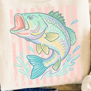 May include: Cream-colored sweatshirt featuring a colorful illustration of a leaping fish. The fish is depicted in shades of blue, green, yellow, and pink, with a red open mouth. Vertical pink stripes are in the background, and water splashes are around the fish.