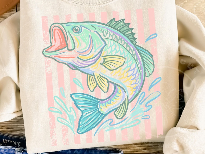 May include: Cream-colored sweatshirt featuring a colorful illustration of a leaping fish. The fish is depicted in shades of blue, green, yellow, and pink, with a red open mouth. Vertical pink stripes are in the background, and water splashes are around the fish.