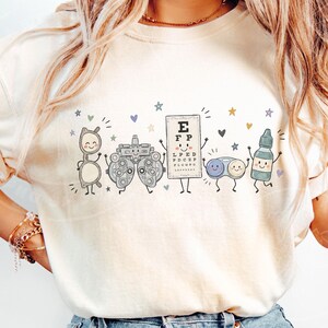 May include: A cream-colored t-shirt featuring a graphic print of five anthropomorphic optometry items: a pair of glasses, a phoropter, an eye chart with the letters EFP, a contact lens case, and an eye drop bottle, all surrounded by small stars and hearts.