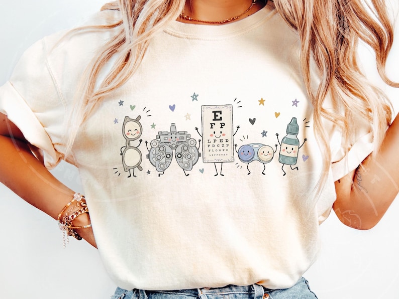 May include: A cream-colored t-shirt featuring a graphic print of five anthropomorphic optometry items: a pair of glasses, a phoropter, an eye chart with the letters EFP, a contact lens case, and an eye drop bottle, all surrounded by small stars and hearts.