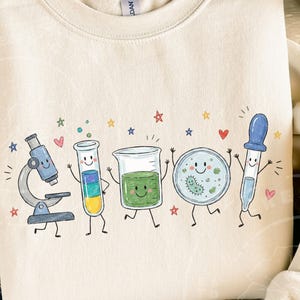 May include: A cream-coloured crewneck sweatshirt featuring a colourful graphic print of five anthropomorphic laboratory items including a microscope, a test tube, a beaker, a petri dish, and a dropper, all decorated with small stars and hearts.