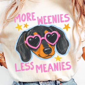 Könnte beinhalten: Cremefarbenes T-Shirt mit einem Cartoon-Dackel, der eine rosa herzförmige Sonnenbrille trägt. Das Shirt hat den Text "MORE WEENIES LESS MEANIES" in Pink, mit gelben Sternakzenten. Ein lustiges, verspieltes Design.