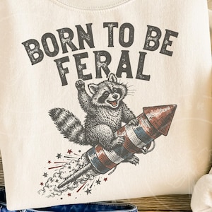 May include: Cream-colored sweatshirt with the text "BORN TO BE FERAL" above a raccoon riding a rocket. The raccoon is gray and white, and the rocket is red, white, and blue. Stars and streaks of smoke emanate from the rocket.