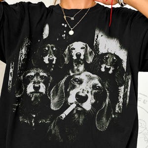 Puede incluir: Una camiseta negra de manga corta con un gran estampado gráfico blanco de alto contraste de cinco perros salchicha. Un perro en primer plano aparece con un cigarrillo en la boca. El diseño tiene una estética desgastada de estilo vintage.