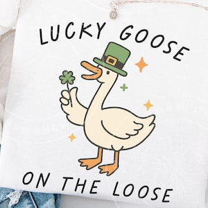 May include: White sweatshirt featuring a cartoon goose wearing a green hat and holding a four-leaf clover. The text "LUCKY GOOSE ON THE LOOSE" is printed above and below the goose illustration. The design includes orange stars and a green cross.