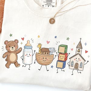 May include: A cream-colored t-shirt featuring a colorful graphic print of a brown teddy bear, a baby bottle, a wooden ark with a giraffe and elephant, a stack of three alphabet blocks, and a church, all with smiling faces and small stars.
