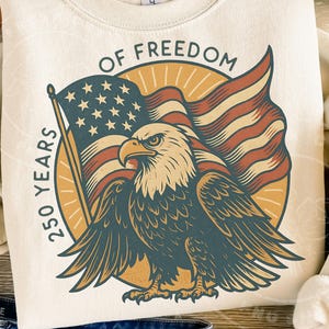 Może przedstawiać: Kremowa bluza z patriotycznym wzorem. Grafika zawiera amerykańską flagę, bielika amerykańskiego oraz tekst "OF FREEDOM" i "250 YEARS". Orzeł ma rozłożone skrzydła, a flaga powiewa za nim.