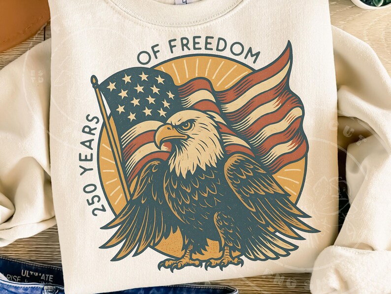 Może przedstawiać: Kremowa bluza z patriotycznym wzorem. Grafika zawiera amerykańską flagę, bielika amerykańskiego oraz tekst "OF FREEDOM" i "250 YEARS". Orzeł ma rozłożone skrzydła, a flaga powiewa za nim.