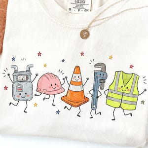 Op de afbeelding: Een crèmekleurig t-shirt met een grafische print van vijf antropomorfe bouw-personages: een grijze gasmeter, een roze veiligheidshelm, een oranje verkeerskegel, een blauwe pijptang en een neongeel veiligheidsvest met reflecterende strepen.