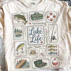 May include: Cream-colored t-shirt featuring a "Lake Life" design. The design includes illustrations of a fish, fishing gear, a boat, a life preserver, and the words "Lake Life" in a decorative font. The shirt has short sleeves and a crew neck.
