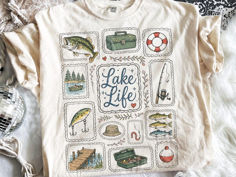 May include: Cream-colored t-shirt featuring a "Lake Life" design. The design includes illustrations of a fish, fishing gear, a boat, a life preserver, and the words "Lake Life" in a decorative font. The shirt has short sleeves and a crew neck.