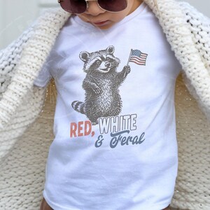 May include: White t-shirt featuring a raccoon holding an American flag. The text "RED, WHITE & FERAL" is printed below the raccoon in red, white, and blue. The shirt is worn by a person wearing a cream-colored cardigan.
