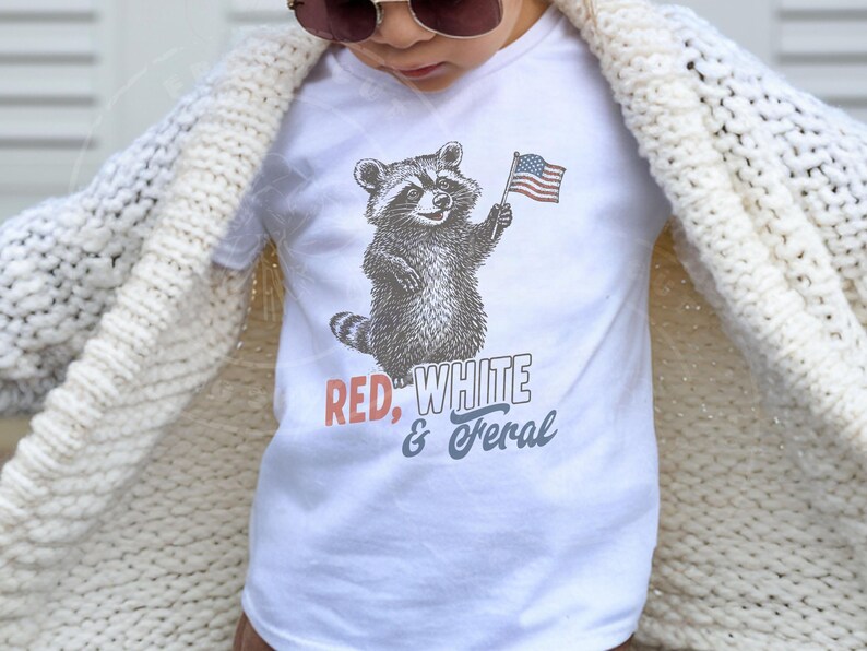May include: White t-shirt featuring a raccoon holding an American flag. The text "RED, WHITE & FERAL" is printed below the raccoon in red, white, and blue. The shirt is worn by a person wearing a cream-colored cardigan.