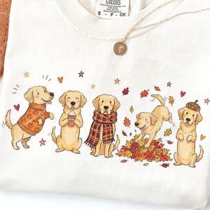 May include: A cream-colored t-shirt featuring a graphic illustration of five golden retriever dogs in autumn-themed attire and activities, including wearing a pumpkin sweater, holding a coffee cup, wearing a plaid scarf, playing in leaves, and wearing an acorn hat.